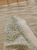 A close-up of a folded baby blanket or swaddle on a bed. The blanket features a cream base with a small green floral pattern on one side and a soft, textured cream grid pattern on the other, with a sage green border.