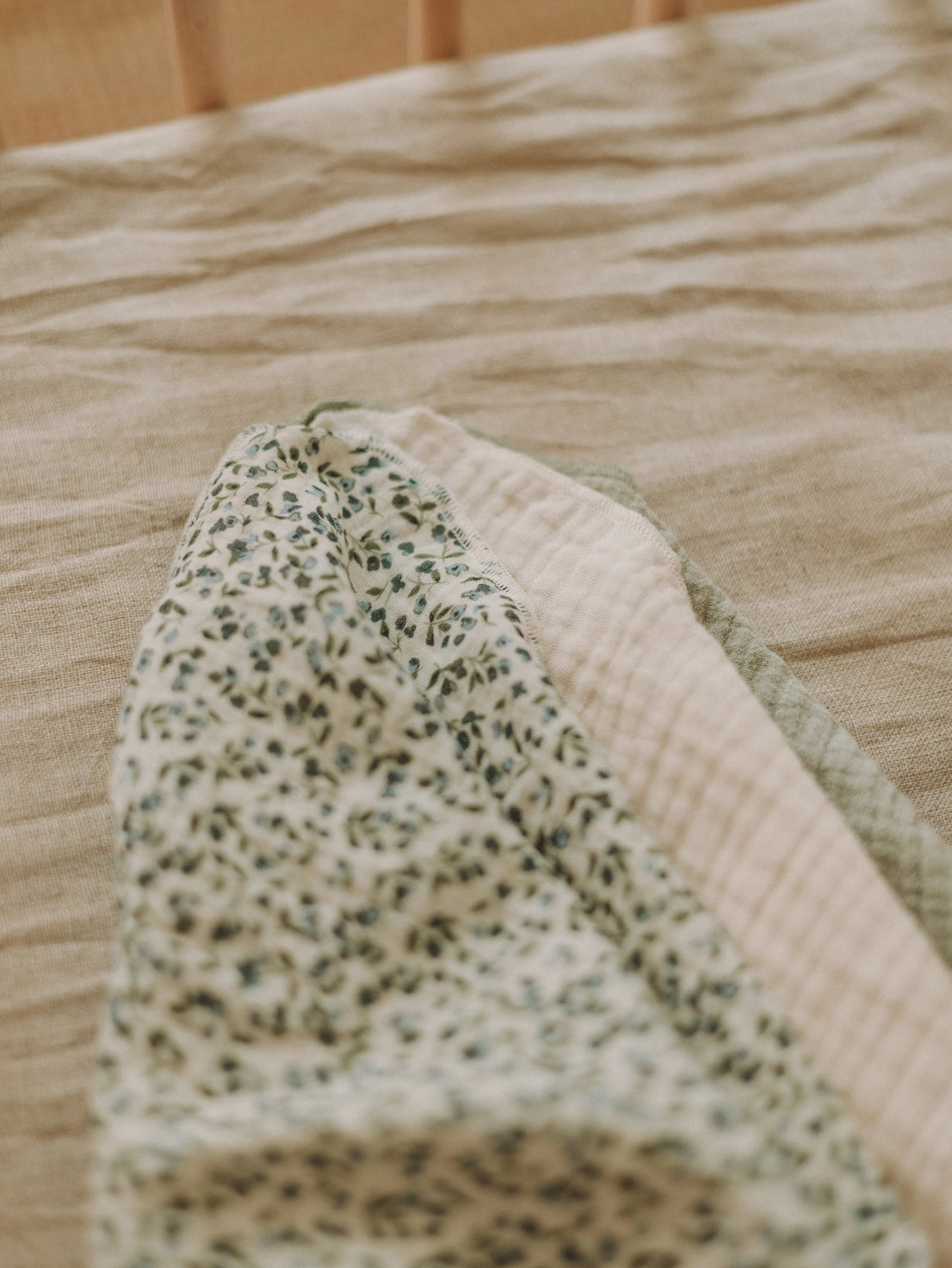 A close-up of a folded baby blanket or swaddle on a bed. The blanket features a cream base with a small green floral pattern on one side and a soft, textured cream grid pattern on the other, with a sage green border.