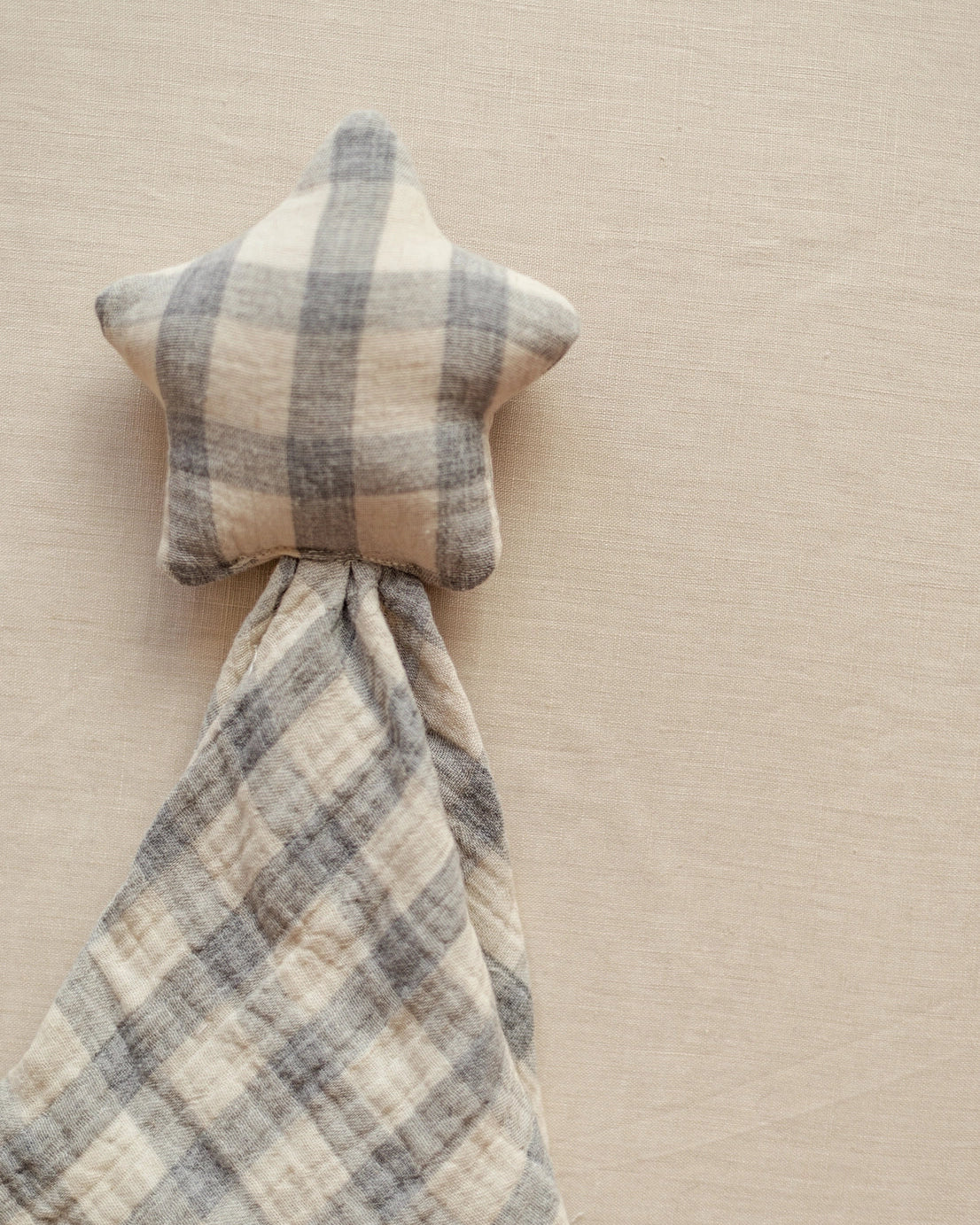A star-shaped baby lovey blanket with a soft plaid pattern in beige and gray, shown on a neutral beige fabric background.