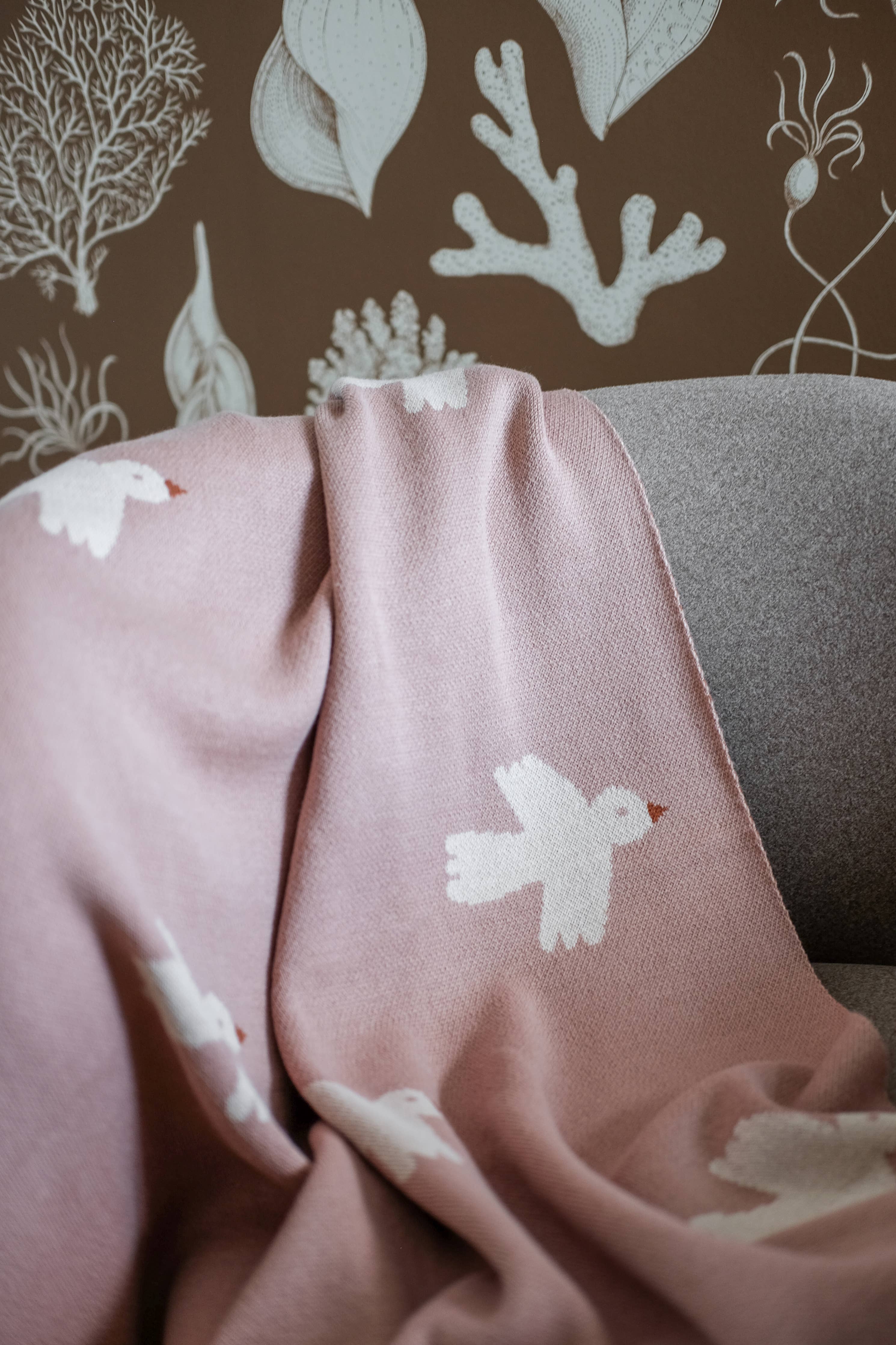 Pink blanket with white bird designs draped over a gray surface against a brown background with white coral and leaf patterns.
