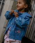 A young child wearing a blue denim jacket with a pink rose embroidery on the pocket, paired with a pink patterned shirt, standing against a rustic wooden wall.