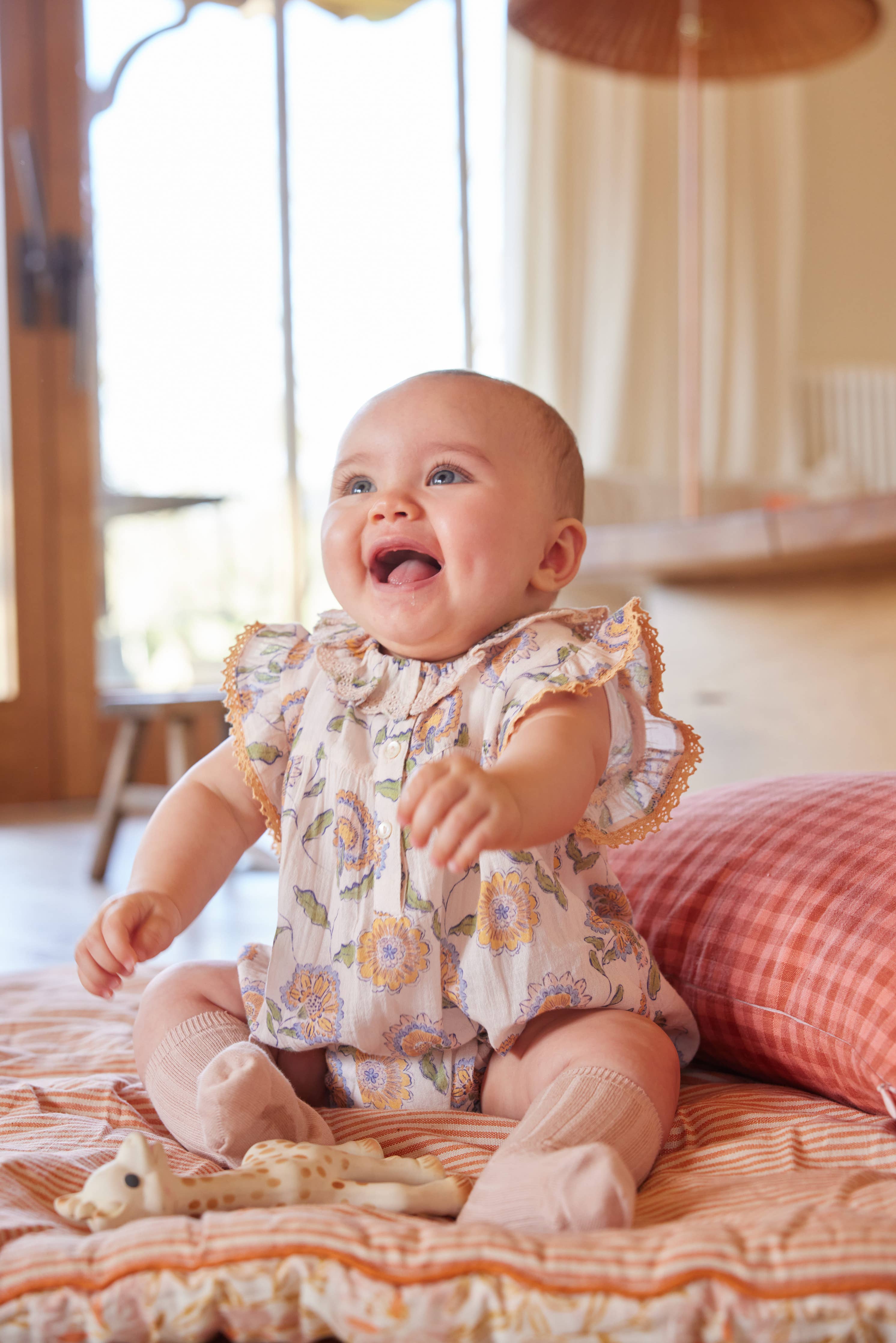 Baby in a floral outfit sitting on a bed with a blurred background