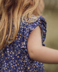 A close-up lifestyle shot of a young girl wearing a blue floral dress, viewed from behind as she looks to the side. The dress features a delicate white and yellow floral pattern on a deep blue background.