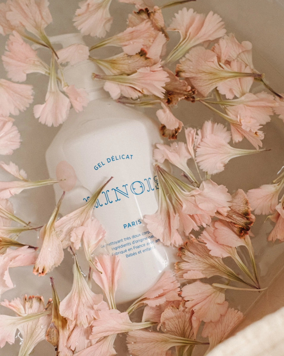 A white bottle of Gel Délicat Minois Paris body wash surrounded by soft pink flowers, set in a warm-toned ceramic bowl.