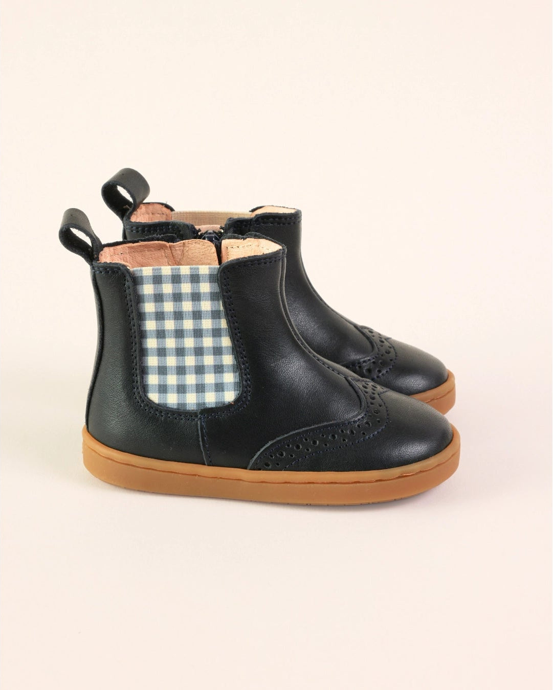 A pair of toddler black leather Chelsea boots with a blue and white gingham elastic side panel and tan soles, shown against a solid pale peach background.
