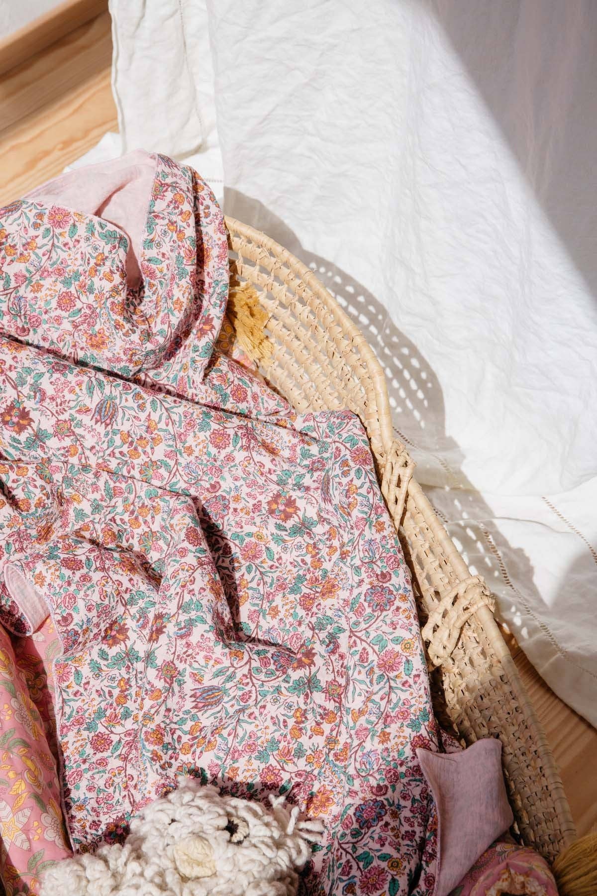 A folded pink floral-patterned fabric garment, likely a robe or blanket, styled in a woven straw basket with a white textured throw blanket on a wooden surface.