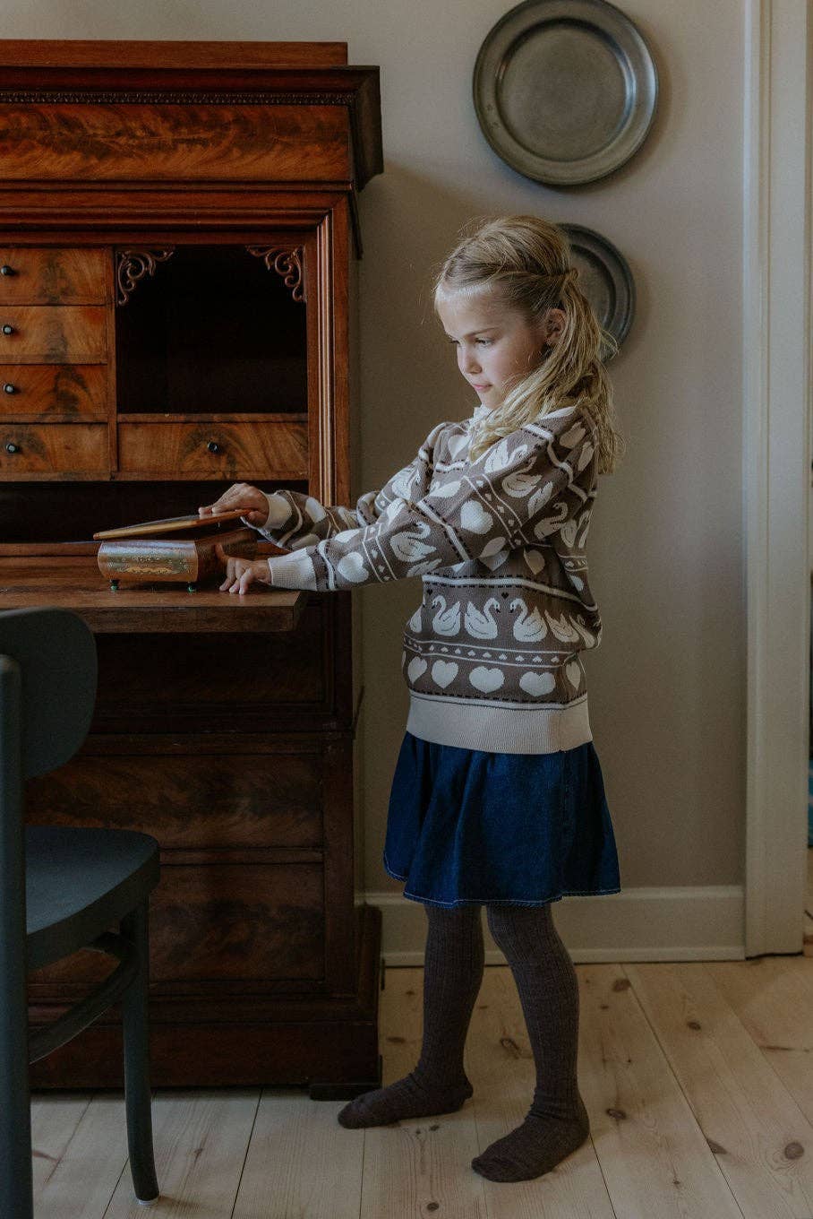A young girl with blonde hair stands in a room next to a dark wooden writing desk, wearing a cream and brown patterned knit sweater, a navy blue skirt, and dark brown socks.