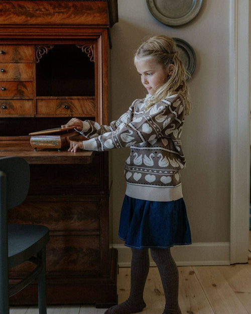A young girl with blonde hair stands in a room next to a dark wooden writing desk, wearing a cream and brown patterned knit sweater, a navy blue skirt, and dark brown socks.
