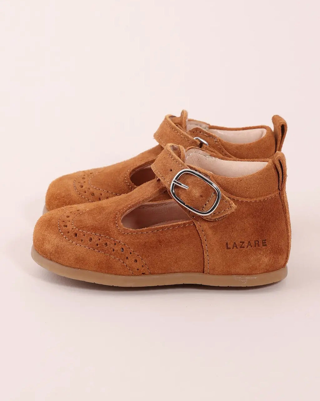 A pair of toddler Mary Jane shoes in tan suede with brogue detailing, featuring a hook-and-loop strap with a silver-tone buckle and the brand name LAZARE printed on the side.