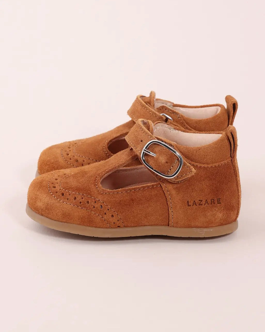 A pair of toddler Mary Jane shoes in tan suede with brogue detailing, featuring a hook-and-loop strap with a silver-tone buckle and the brand name LAZARE printed on the side.