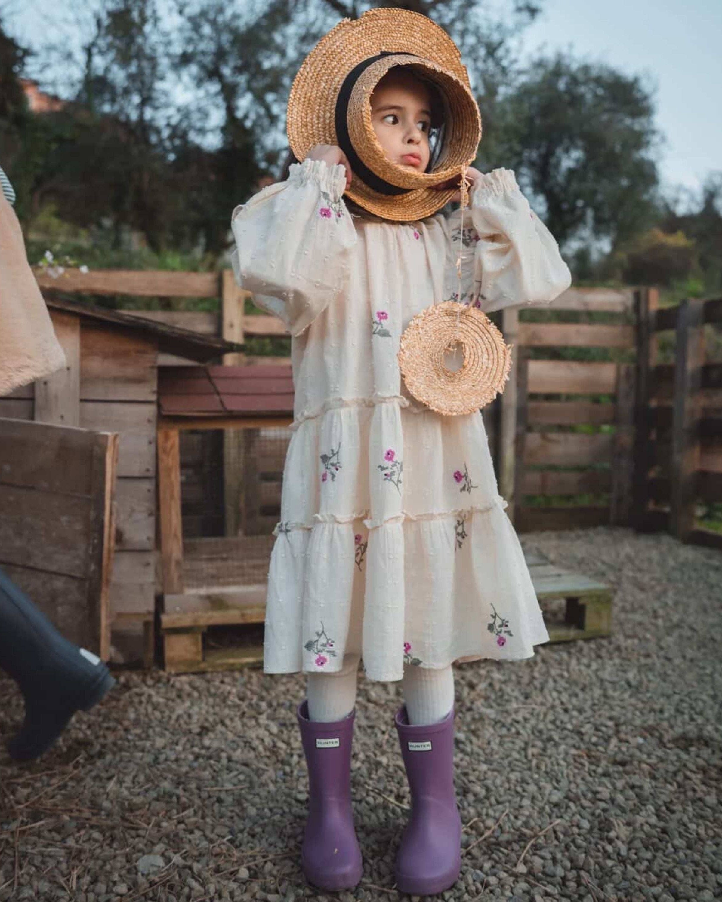 A young girl stands outdoors wearing a cream tiered dress with small floral prints, a large woven straw bonnet, a round woven crossbody bag, and purple rain boots.