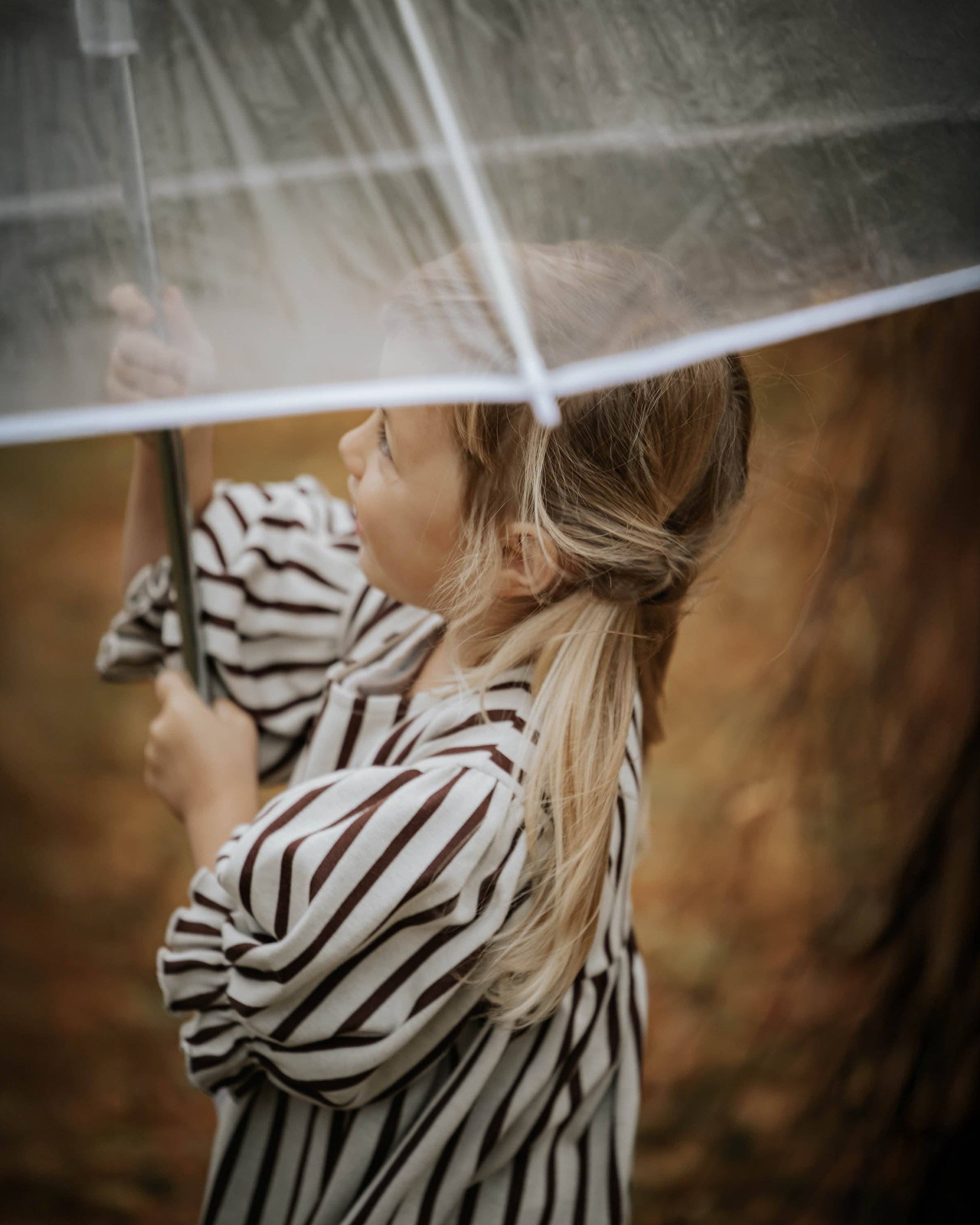 A young girl with blonde hair in a ponytail, wearing a black and white striped long-sleeve top, holding a clear plastic umbrella in a rainy outdoor setting.
