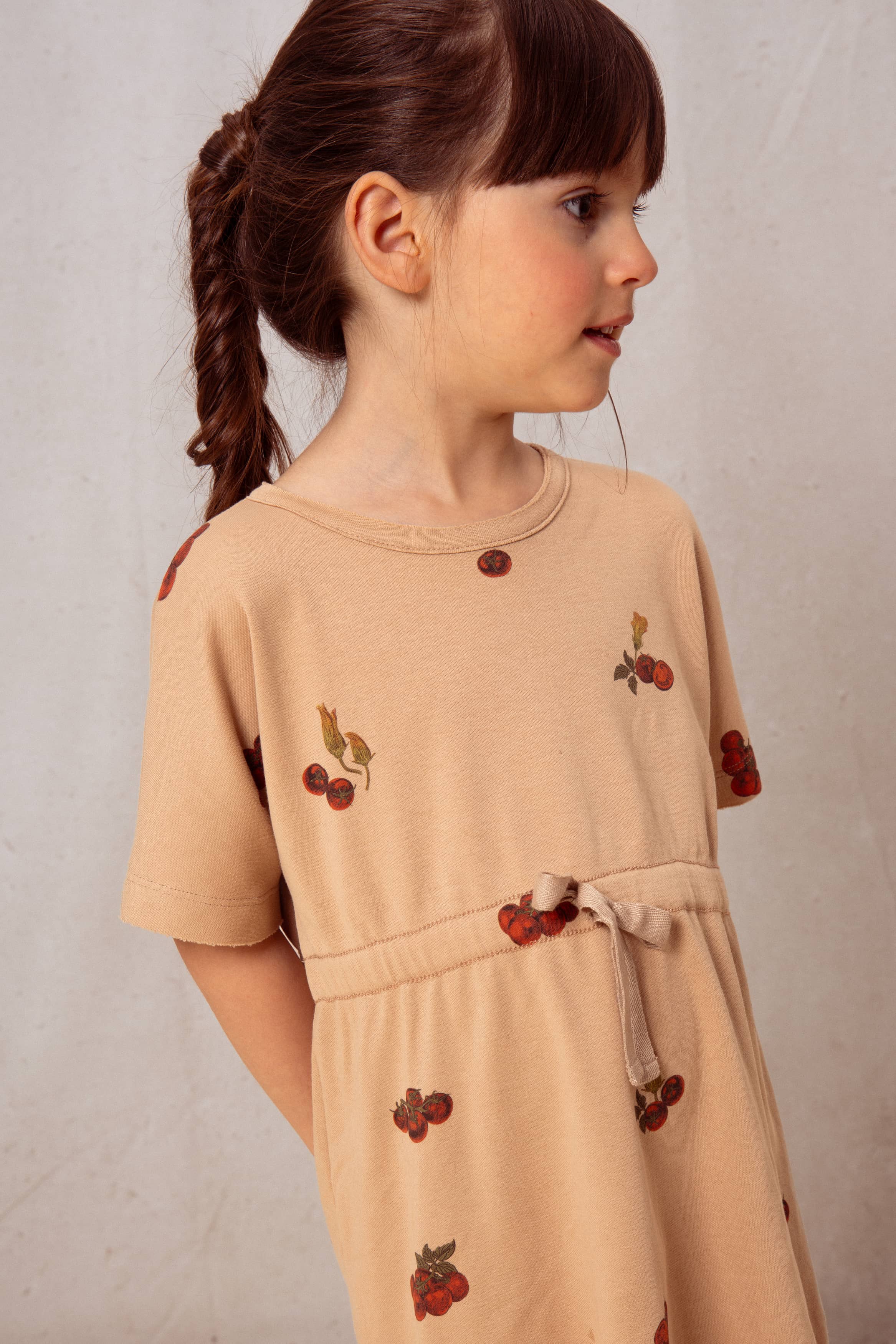 Young girl wearing a beige dress with cherry appliqués against a plain background