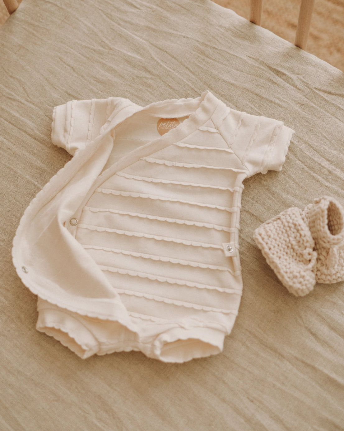 A cream-colored short-sleeve baby bodysuit with horizontal textured stripes and a snap closure, laid flat on a beige linen surface next to a matching knit bootie.