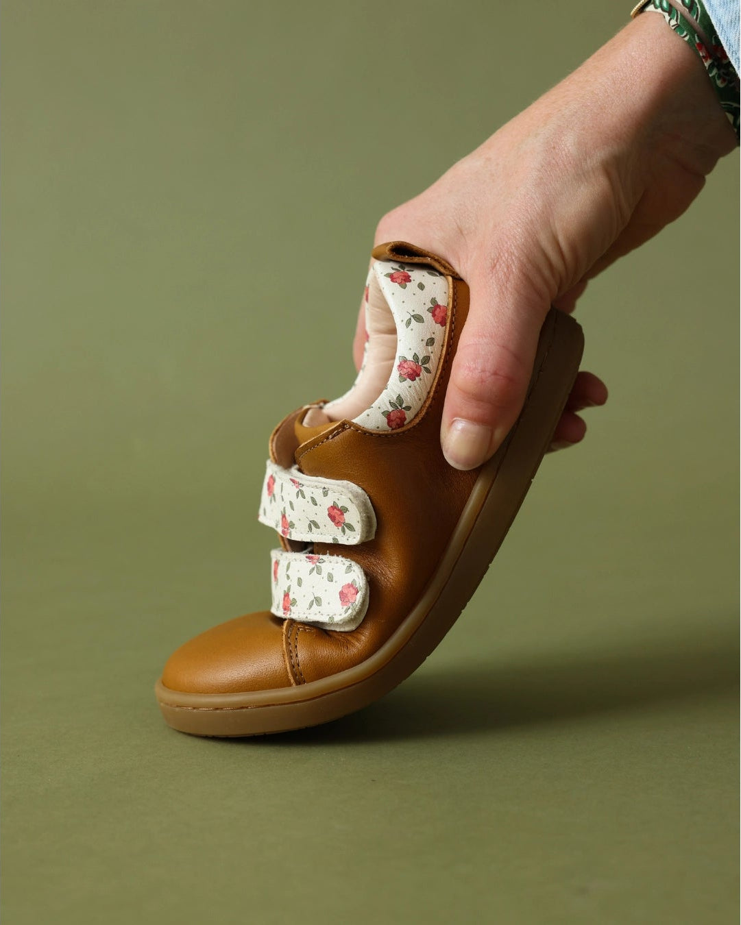 A hand holds a brown leather baby sandal with two adjustable floral-patterned velcro straps against a solid olive green background.