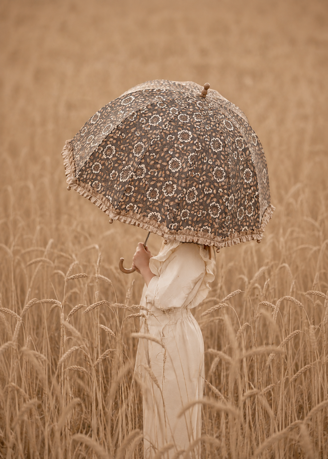 A person in a long cream dress holding a lace parasol umbrella with fringe, standing in a golden wheat field.