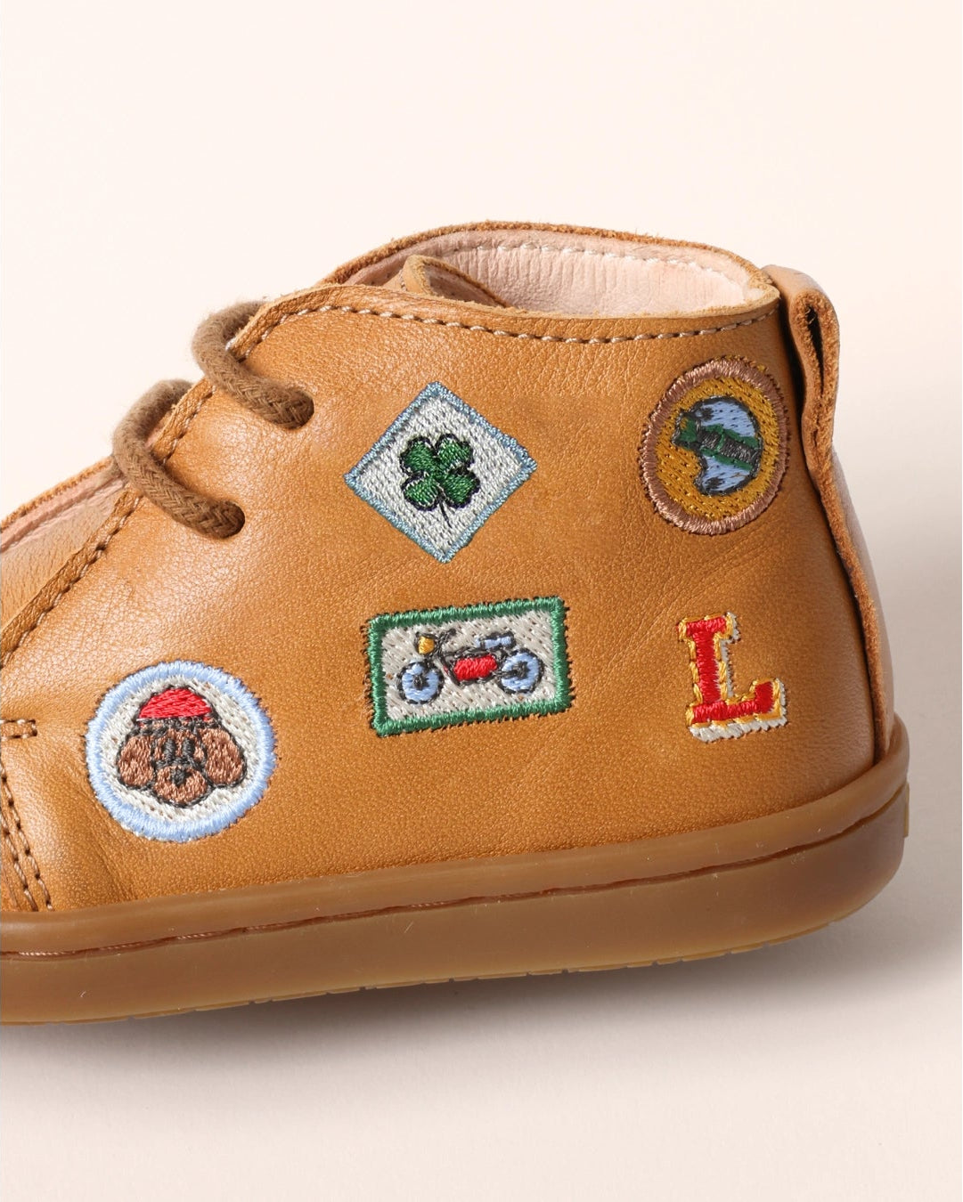 A close-up of a tan leather baby bootie decorated with colorful embroidered patches, including a green shamrock, a red motorcycle, a brown mushroom, and a red letter 'L'.