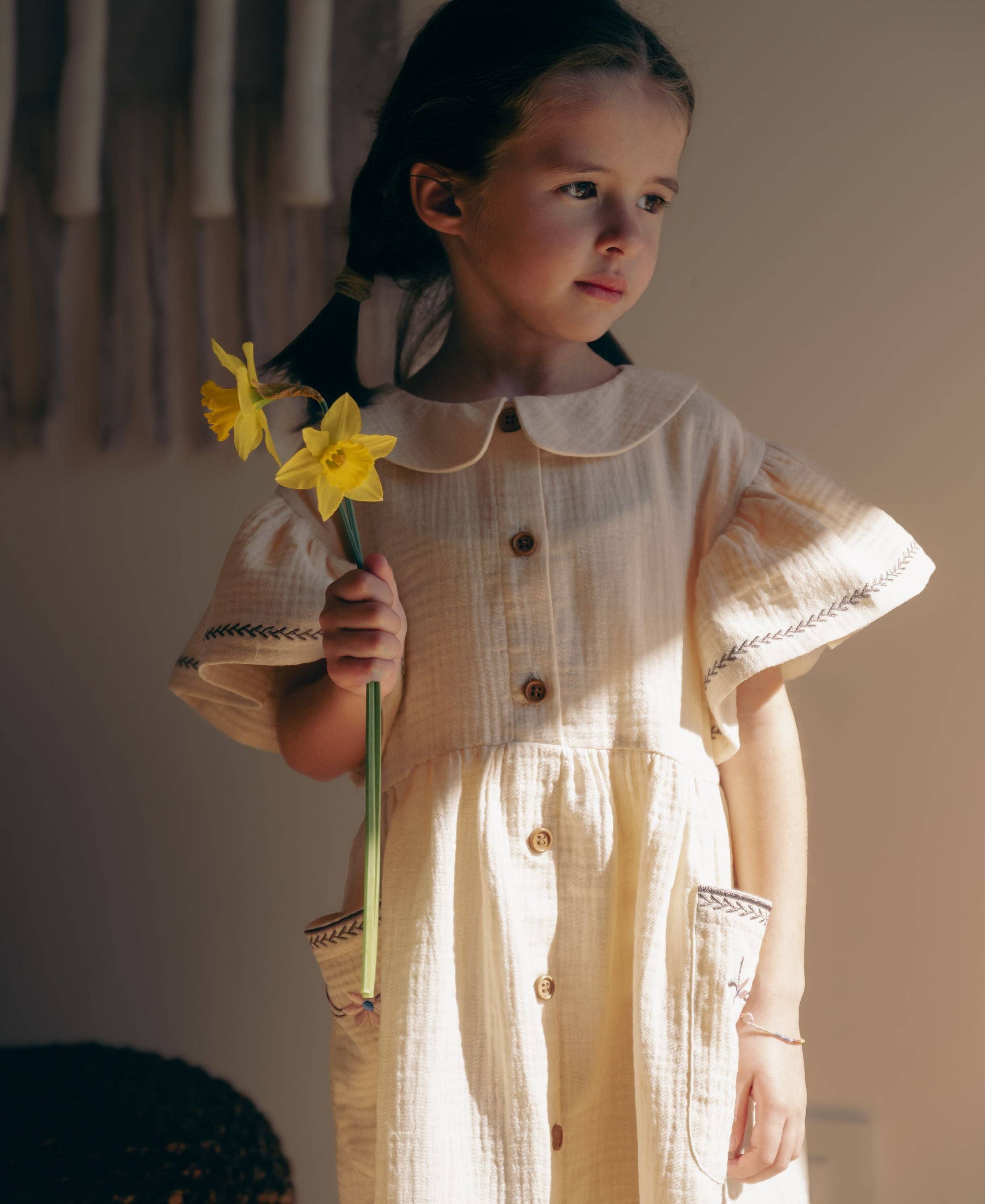 A young girl with dark hair in a ponytail stands indoors holding two yellow daffodils. She is wearing a cream-colored textured cotton dress with a Peter Pan collar, short flutter sleeves with embroidered trim, and front buttons.