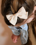 A close-up lifestyle shot of a young child wearing a large, cream-colored fabric bow hair clip. The bow is styled on the child's brown hair, and the child is wearing a light blue denim shirt and a brown dress against a soft, warm beige background.