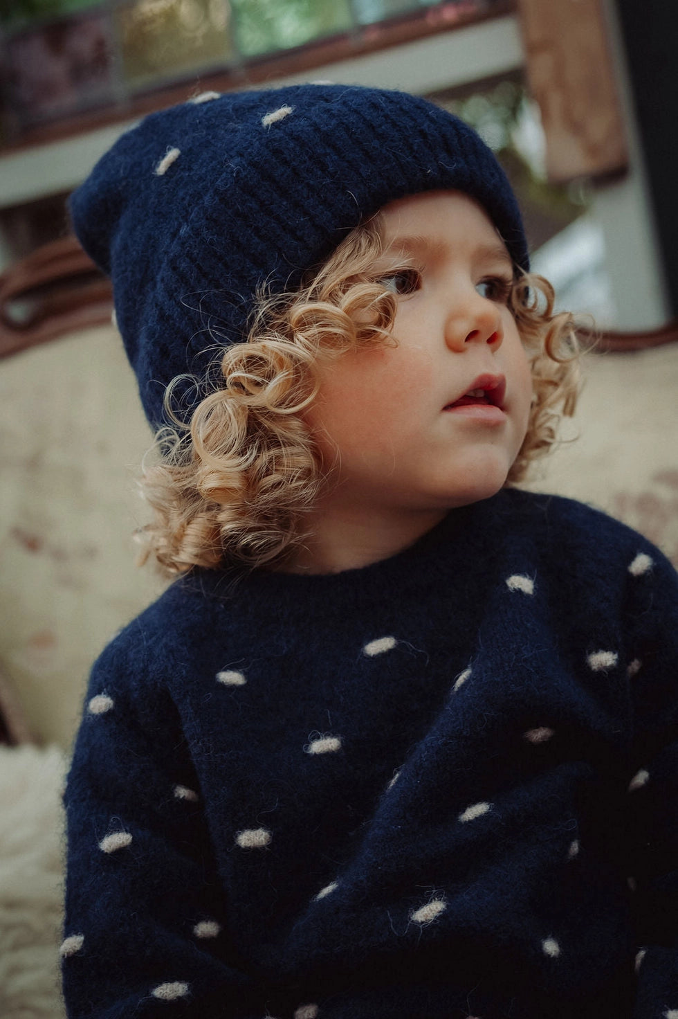 A close-up lifestyle shot of a young child wearing a navy blue knit beanie and matching knit sweater, both decorated with small white polka dots. The child has curly blonde hair and is looking to the side, seated in a cozy indoor setting.