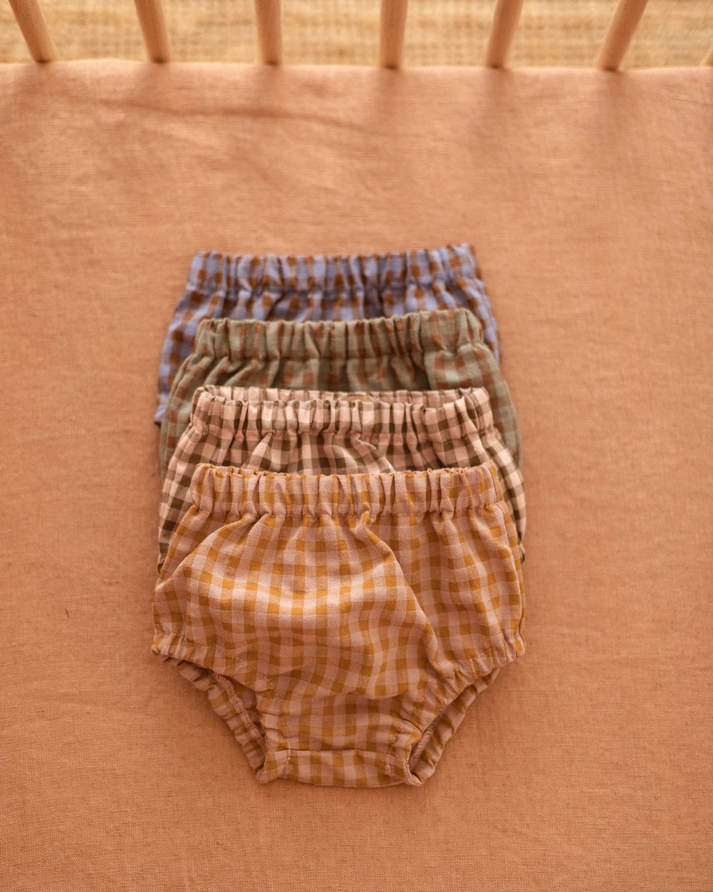 Four pairs of baby bloomers in a soft gingham check pattern, stacked neatly on a peach-colored fabric surface. The set includes blue, green, tan, and mustard yellow checks.