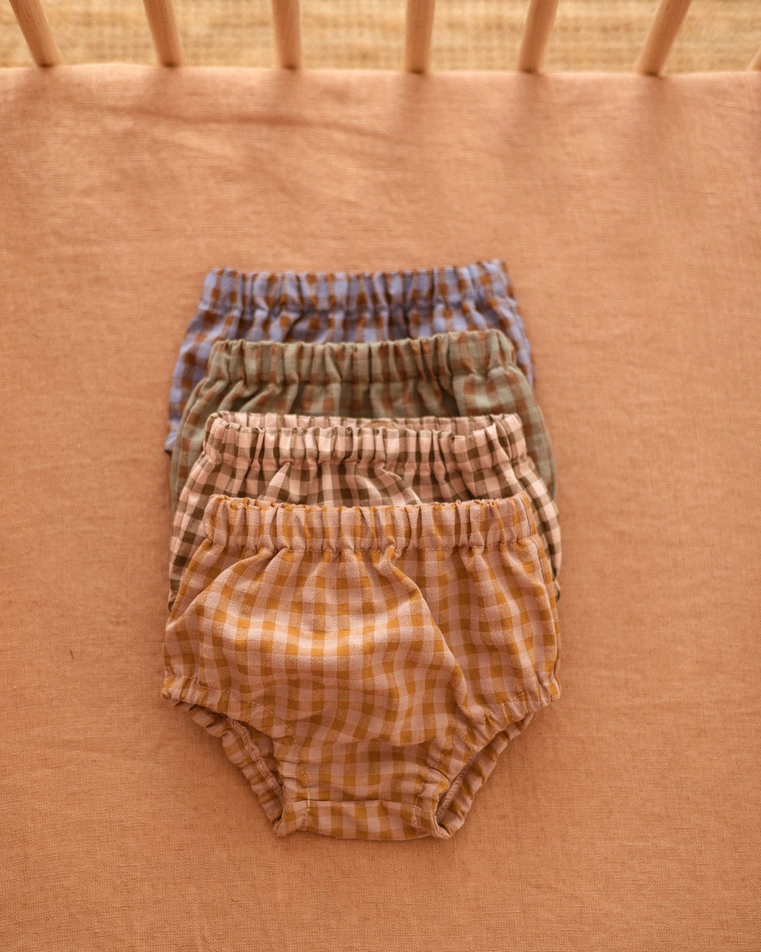 Four pairs of baby bloomers in a soft gingham check pattern, stacked neatly on a peach-colored fabric surface. The set includes blue, green, tan, and mustard yellow checks.