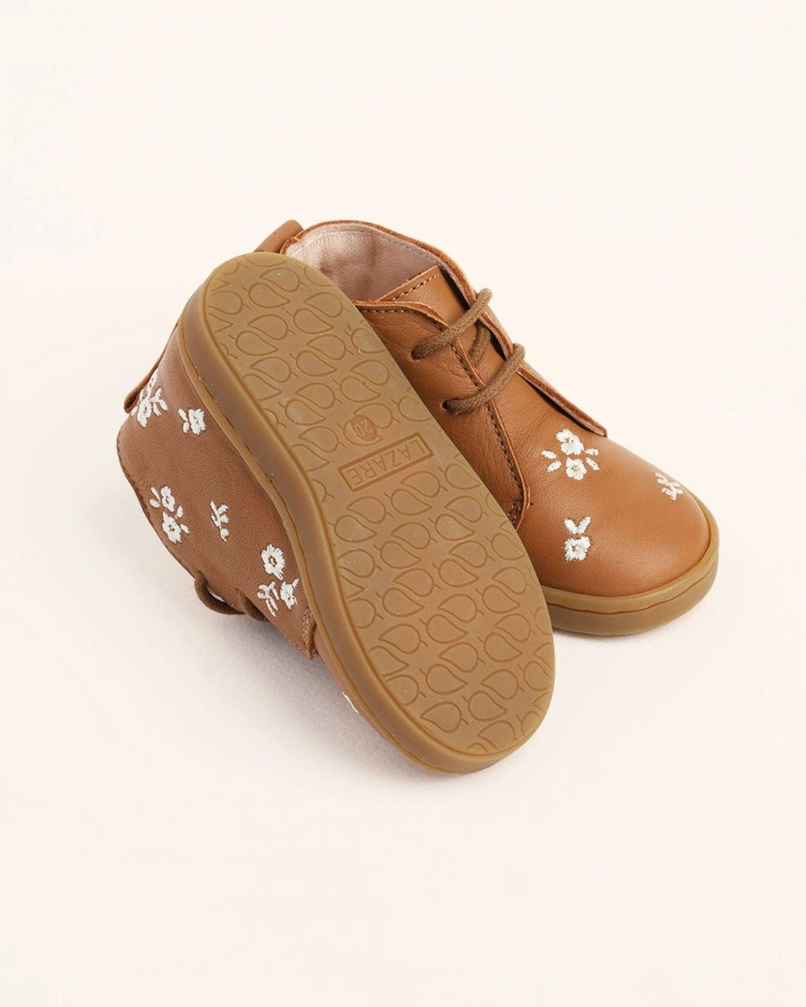A pair of tan leather baby boots with white floral embroidery and a brown textured sole, shown on a clean off-white background.