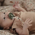 A close-up lifestyle shot of a baby lying on patterned bedding, wearing a beige knit romper and sucking a green pacifier.