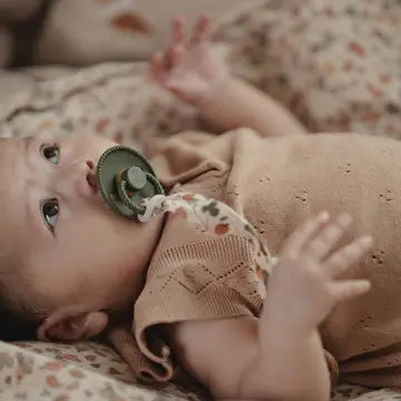 A close-up lifestyle shot of a baby lying on patterned bedding, wearing a beige knit romper and sucking a green pacifier.