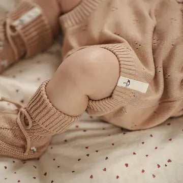 A close-up of a baby's legs wearing a beige knit romper and matching knit booties, lying on a heart-patterned blanket.
