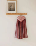 A dusty rose velvet cape with a matching hood hangs from a wooden coat rack mounted on a plain off-white wall. A small framed print is also hanging above the rack.