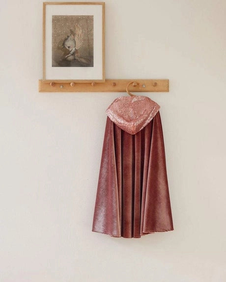 A dusty rose velvet cape with a matching hood hangs from a wooden coat rack mounted on a plain off-white wall. A small framed print is also hanging above the rack.