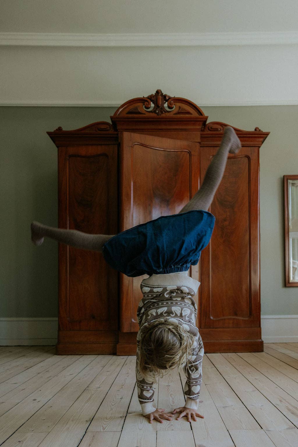 A child wearing a dark blue skirt and patterned top does a handstand on a light wood floor in front of a large dark wood armoire.