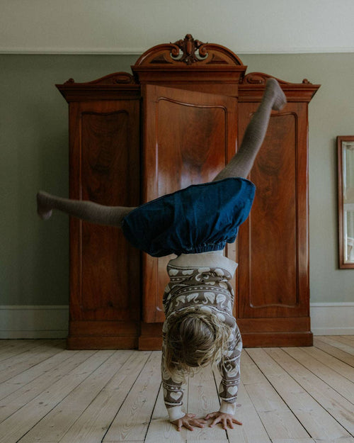 A child wearing a dark blue skirt and patterned top does a handstand on a light wood floor in front of a large dark wood armoire.