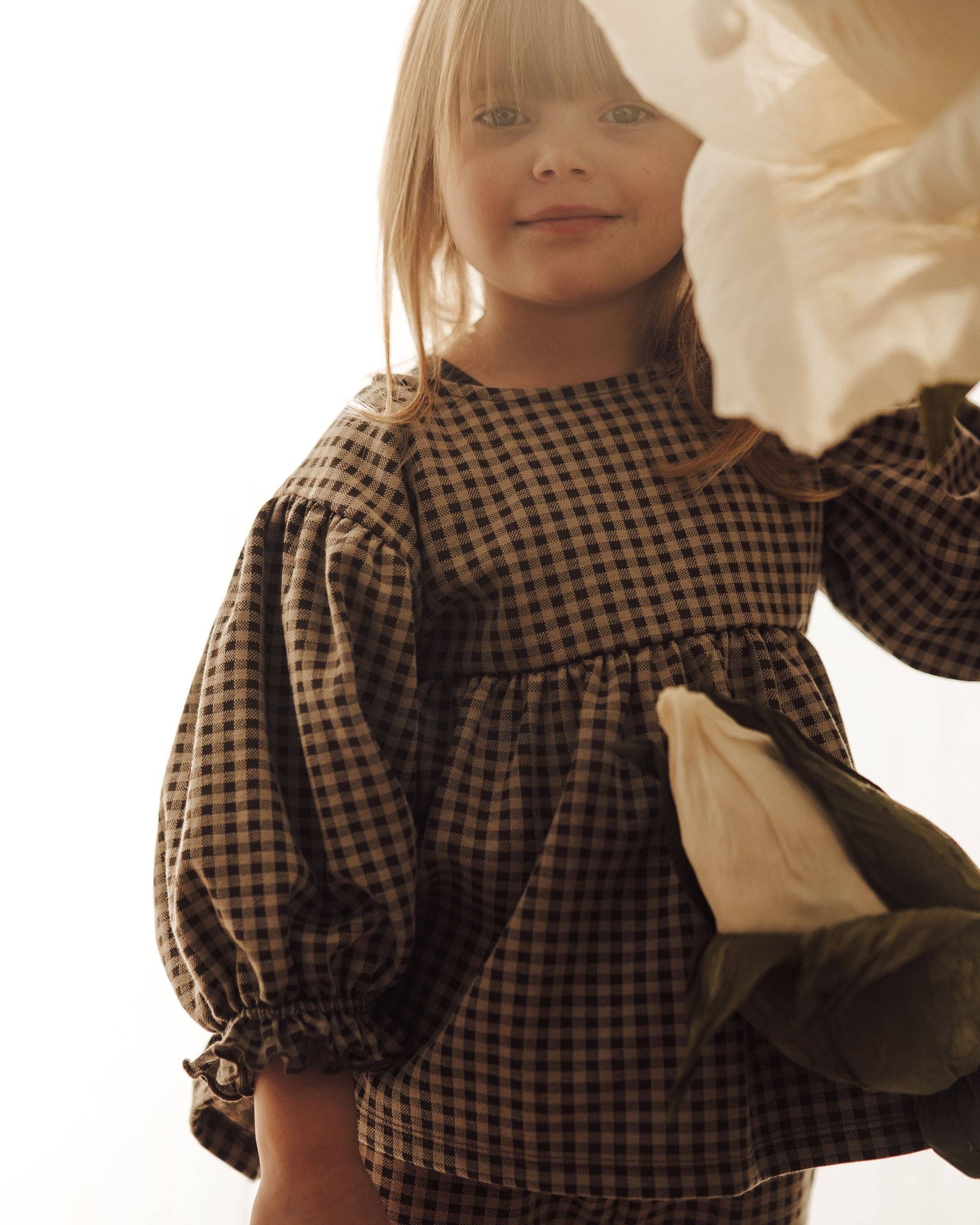 A young girl wearing a brown and black gingham long-sleeve dress with a gathered waist, holding a cream fabric flower against a bright white background.