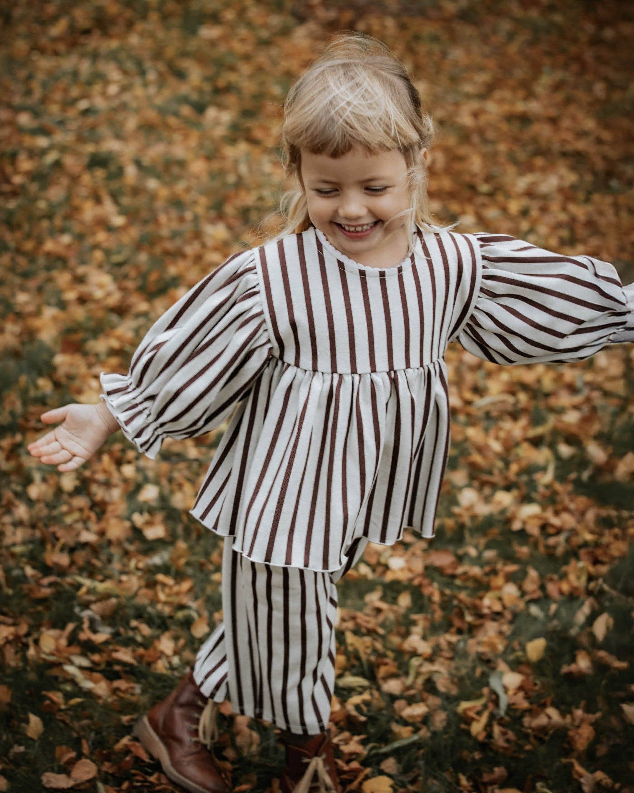 A young child wearing a brown and white vertically striped long-sleeve tunic top and matching striped pants, standing outdoors on a ground covered with fallen autumn leaves.