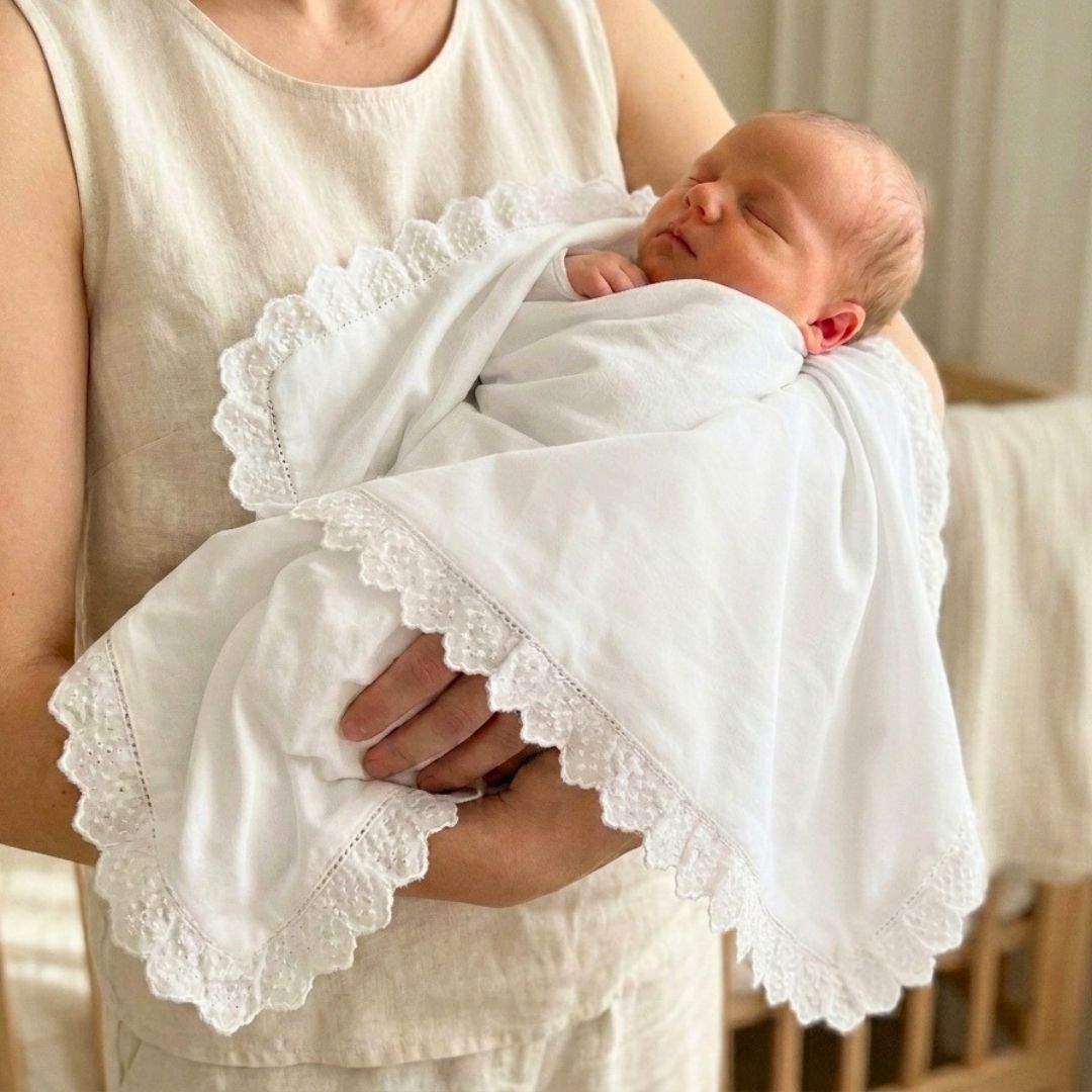 Person holding a baby wrapped in a white blanket with lace trim.