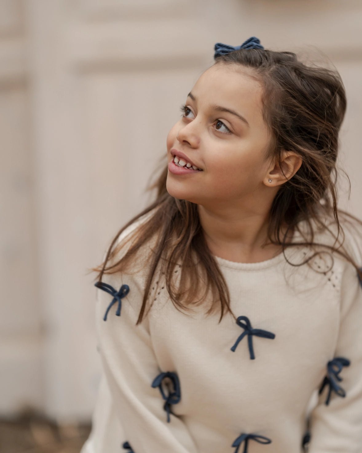 Young girl wearing a beige sweater with blue bows, looking to the side.