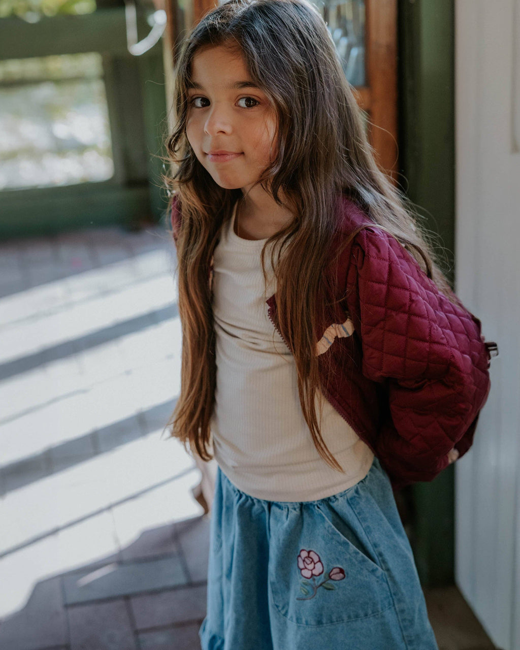 A young girl with long brown hair is shown wearing a cream ribbed t-shirt, a dusty blue denim skirt with a pink flower embroidery, and a burgundy quilted puffer jacket. She is standing in a doorway with natural light casting shadows.