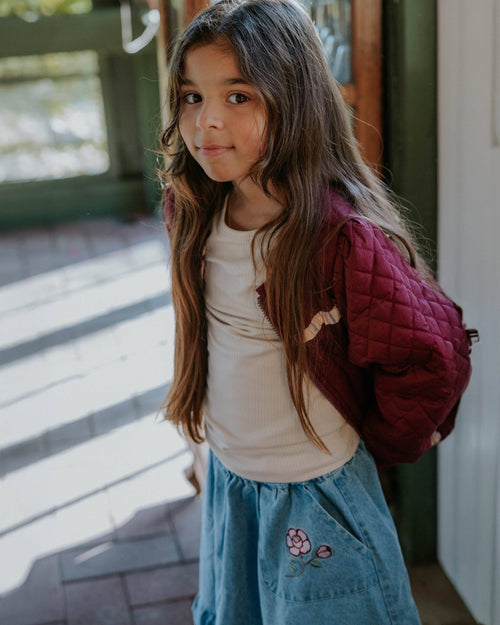 A young girl with long brown hair is shown wearing a cream ribbed t-shirt, a dusty blue denim skirt with a pink flower embroidery, and a burgundy quilted puffer jacket. She is standing in a doorway with natural light casting shadows.