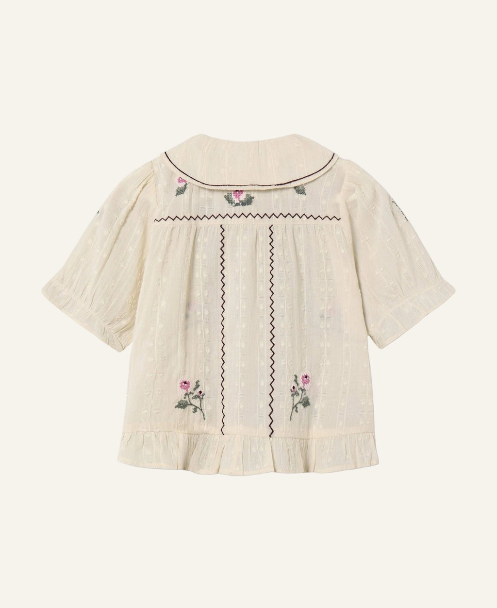 An off-white textured short-sleeve blouse with dark brown zigzag embroidery and pink floral accents, shown from the back on a light gray background.