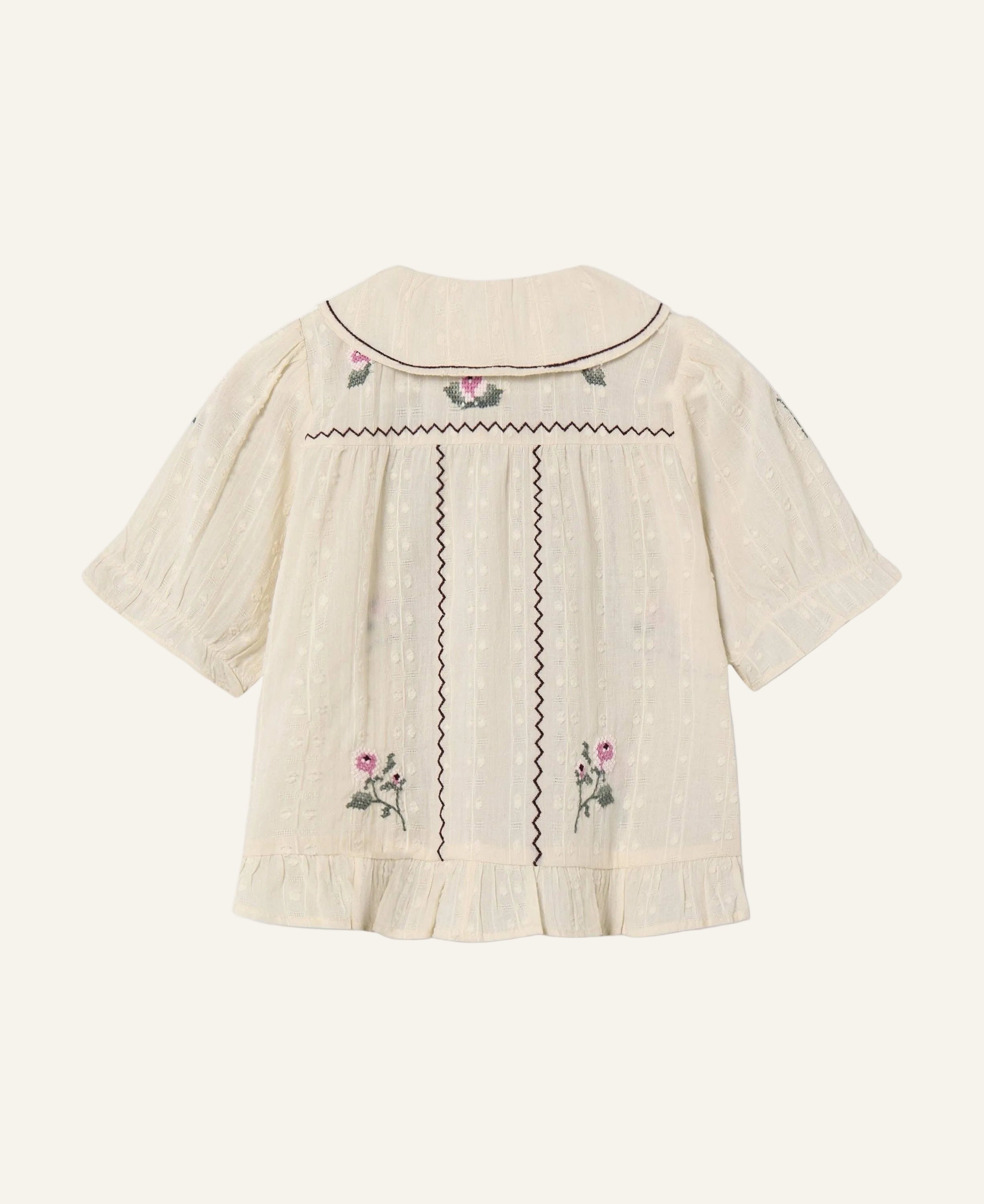 An off-white textured short-sleeve blouse with dark brown zigzag embroidery and pink floral accents, shown from the back on a light gray background.