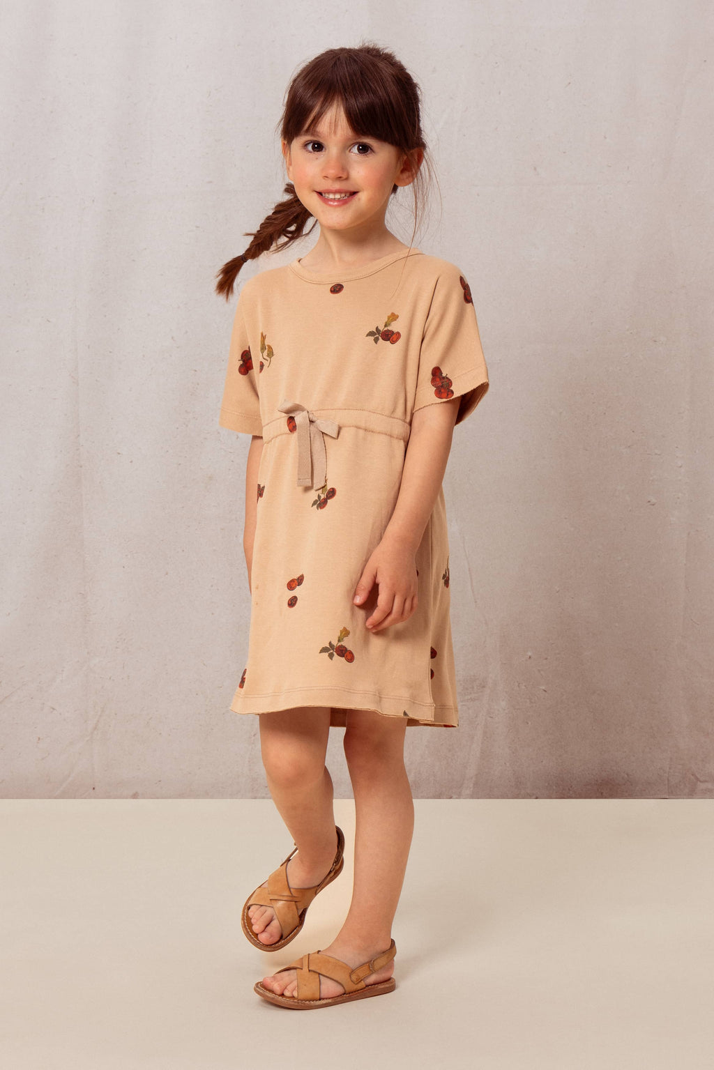 Young girl wearing a beige dress with cherry pattern against a plain background