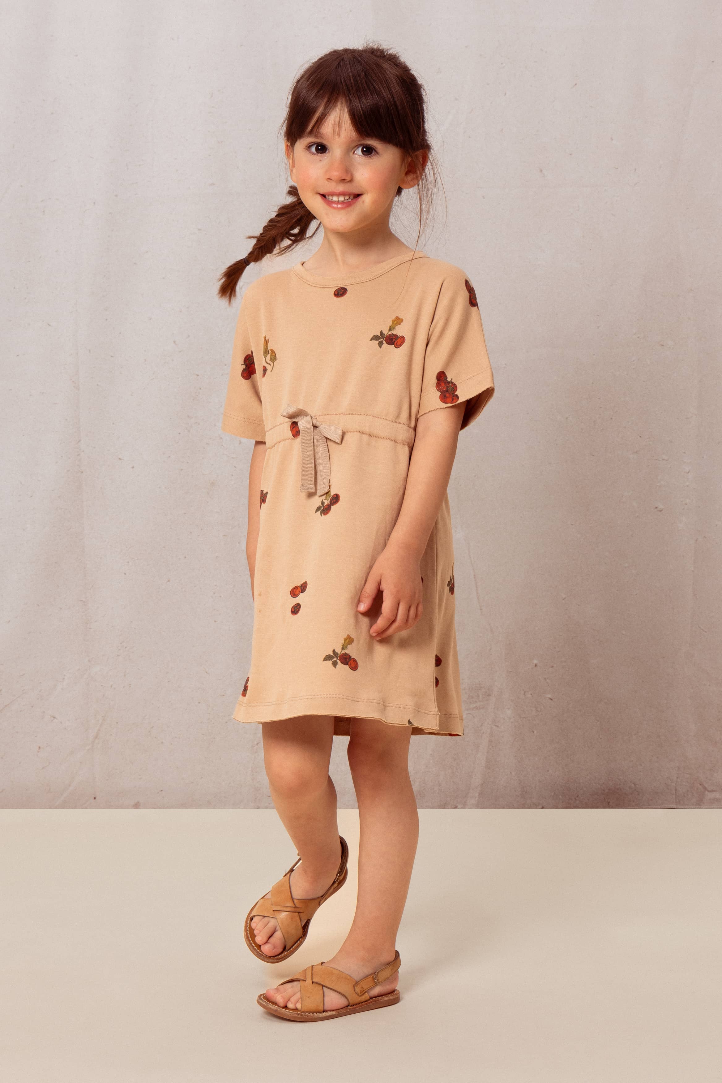 Young girl wearing a beige dress with cherry pattern against a plain background
