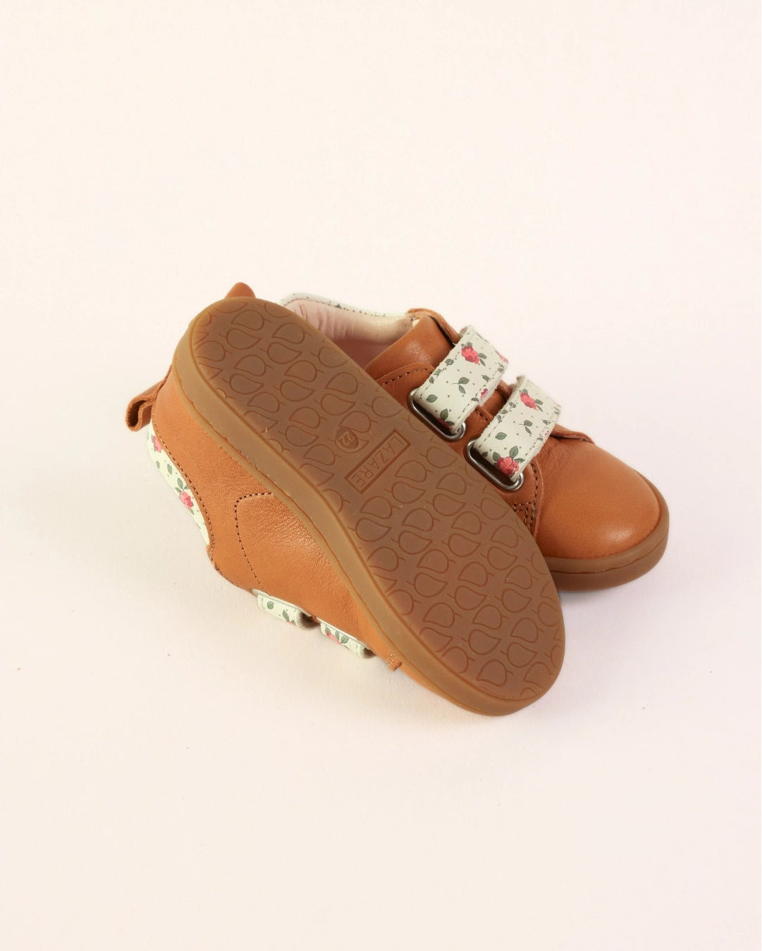 A pair of toddler slip-on shoes in tan leather with a floral-patterned hook-and-loop strap, shown on a plain off-white background.