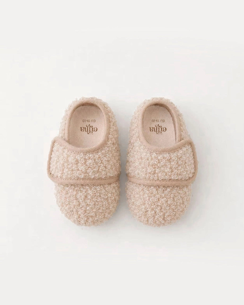 A pair of soft, textured baby slipper moccasins in a warm beige color, shown from a top-down view on a clean white background. The brand name 'ellaa' is visible on the insole of each slipper.