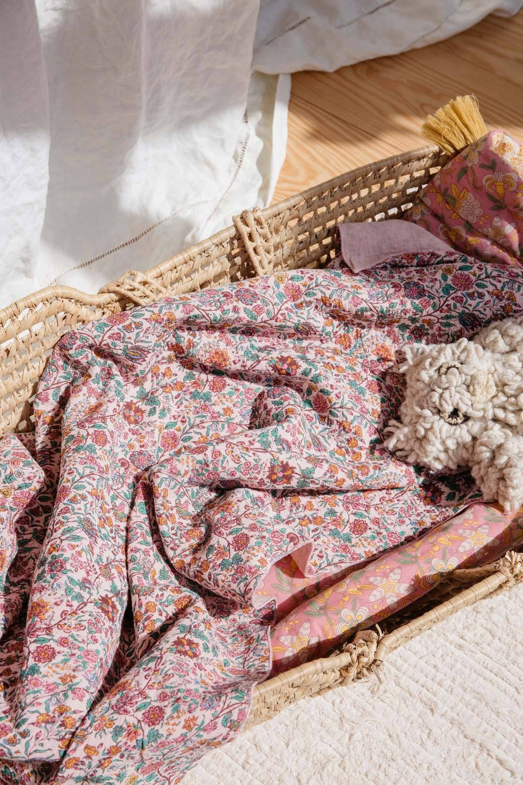 A close-up of a woven rattan basket containing a pink floral patterned blanket or throw, with a small plush toy and folded fabric pieces inside. The scene is styled on a light surface with warm natural light and a wooden floor in the background.