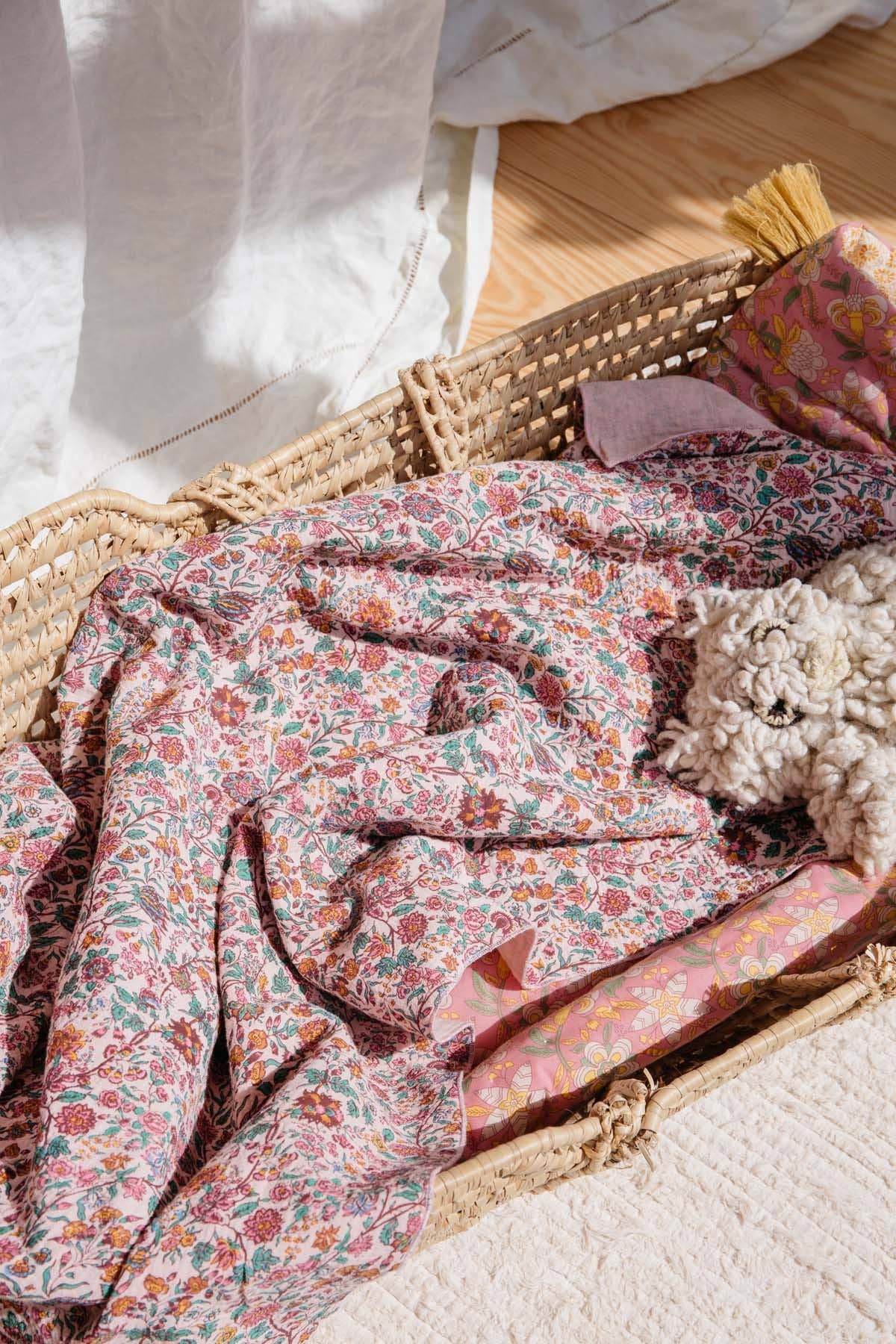 A close-up of a woven rattan basket containing a pink floral patterned blanket or throw, with a small plush toy and folded fabric pieces inside. The scene is styled on a light surface with warm natural light and a wooden floor in the background.