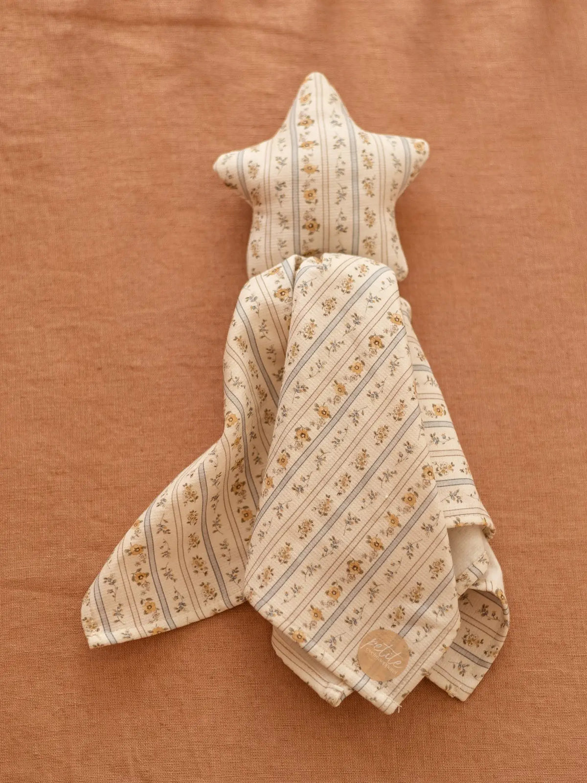A baby teething toy made from a star-shaped plush or stuffed fabric head with a matching fabric body, featuring a delicate cream and tan floral pattern with thin blue stripes, laid flat on a textured terracotta background.