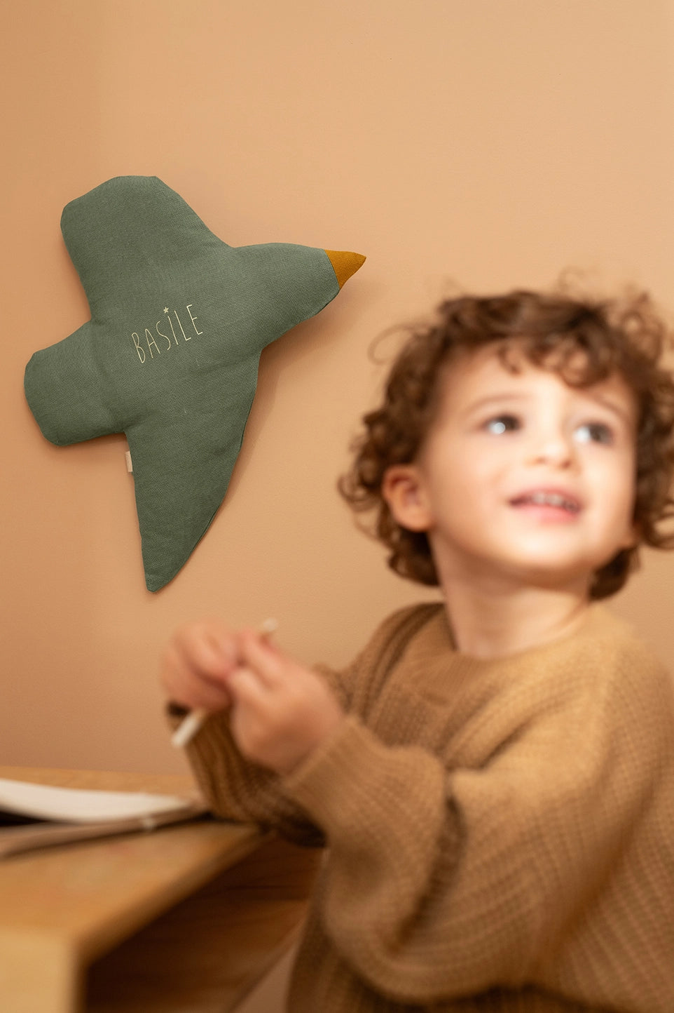 A child wearing a brown knit sweater sits at a wooden table, looking up and smiling. On the beige wall behind them is a green bird-shaped cushion with the name 'BASILE' embroidered in gold thread.